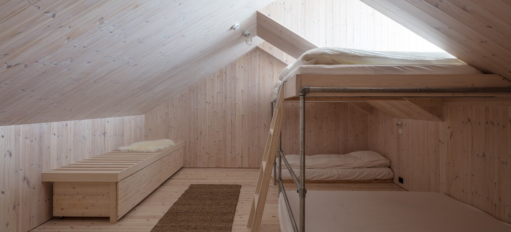 Interiørbilde av et loft med naturlig lys treinnredning, køyeseng og benk – lys kommer inn fra takvindu.