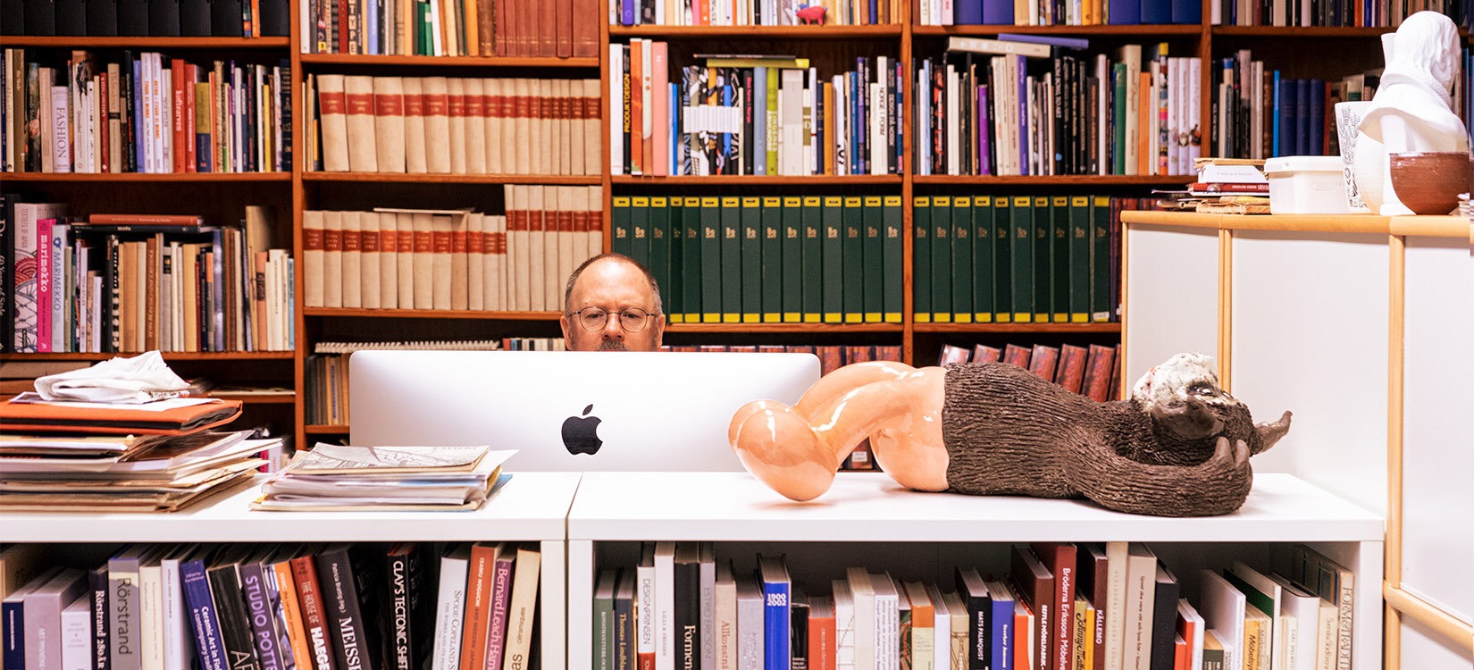 Forfatter og kunsthistoriker Mats Linder foran en mac-skjerm. I forgrunnen og bakgrunnen: bokhyller.