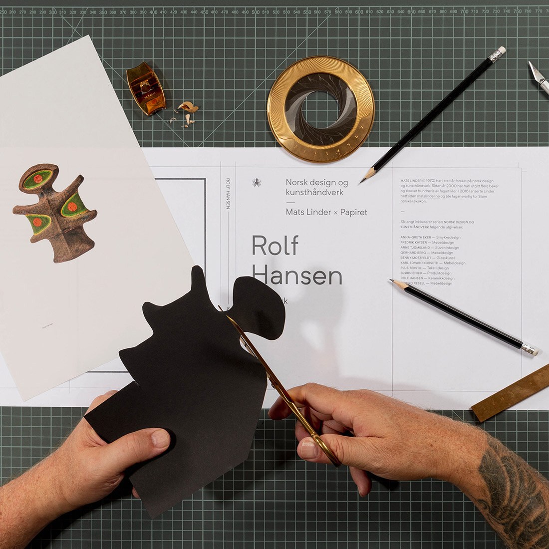 Hånd som jobber ved et bord med åpen bok, blyant, saks og trykksaker.