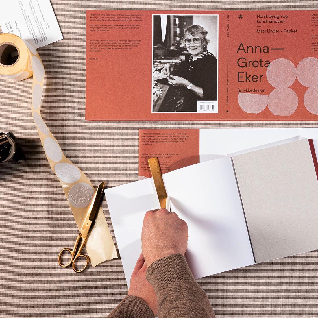 Hender klipper i papir over en arbeidsflate med skisser, tape og blyanter.