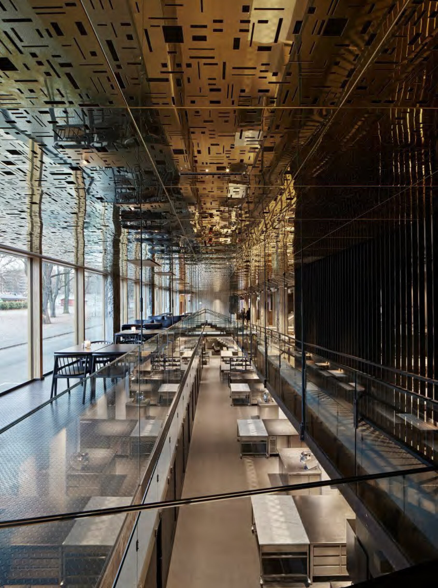 Interiør i moderne bygg med åpne kjøkkenstasjoner i midten, lange arbeidsbenker i stål, glassrekkverk og et dekorativt metalltak, sett fra en øvre etasje.