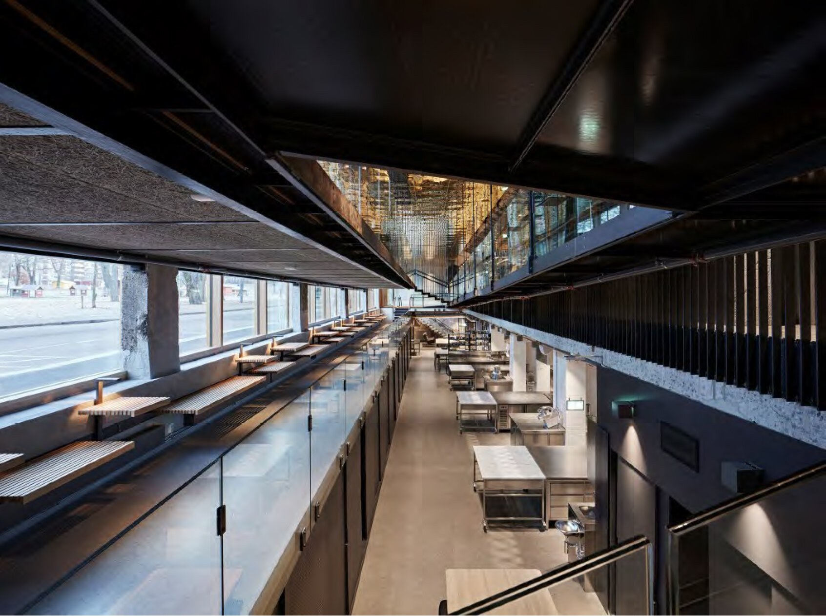 Et interiørfoto som viser et industrielt kjøkken sett fra en messanin med lave benker langs vinduer ut mot gaten. Åpning i taket gir utsyn opp mot øvre etasjer.