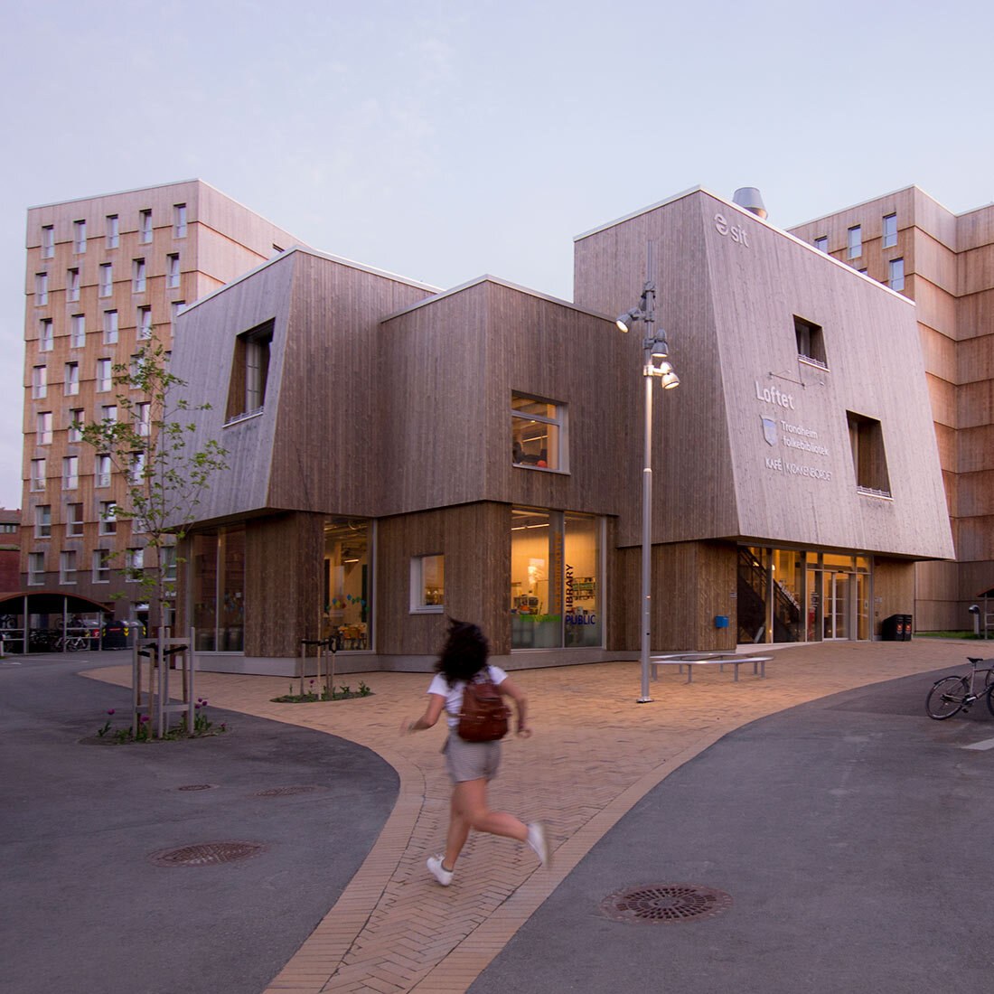 Bildet viser et moderne toetasjes bygg i trekledning med vinklede fasader og en utadrettet førsteetasje. En ung persom løper over plasseni forkant av bygget. I bakgrunnen ses boligblokker i en annen type trefasade.