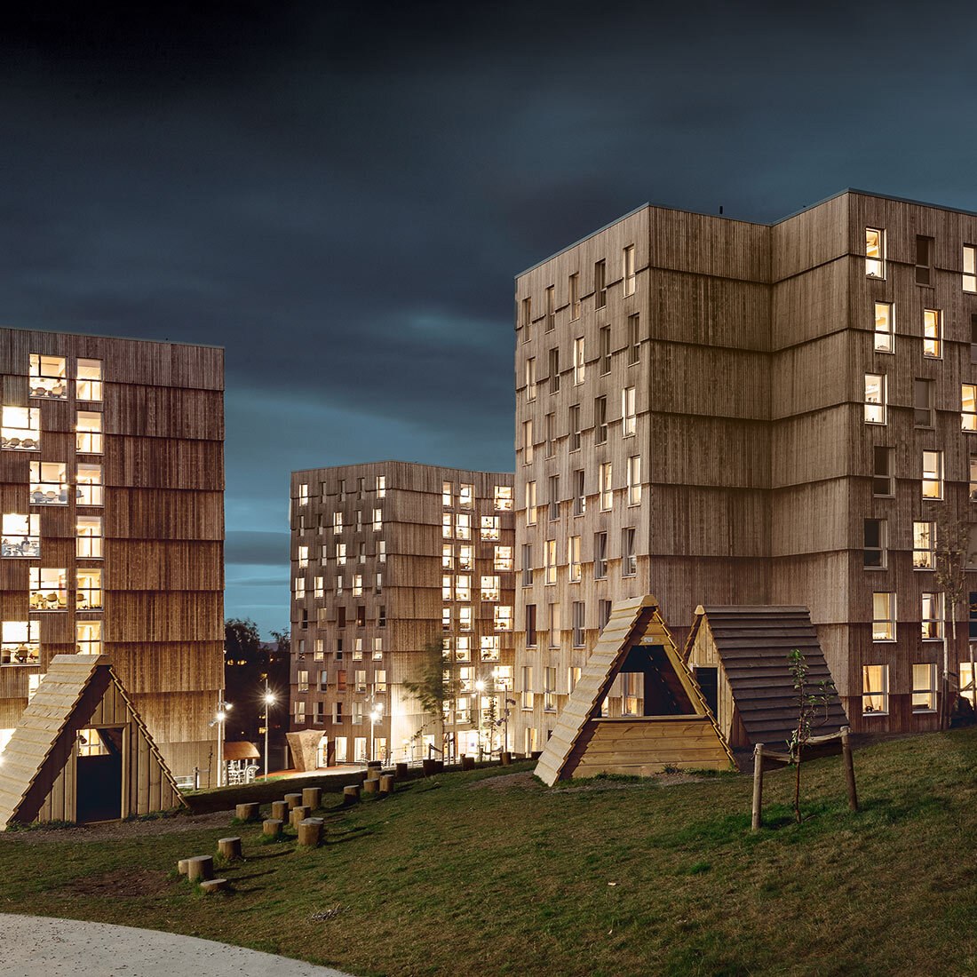 Bildet er tatt nattestid, og viser et lekeområde på en plen i forkant. Deretter flere mellomhøye blokker kledd i tre.