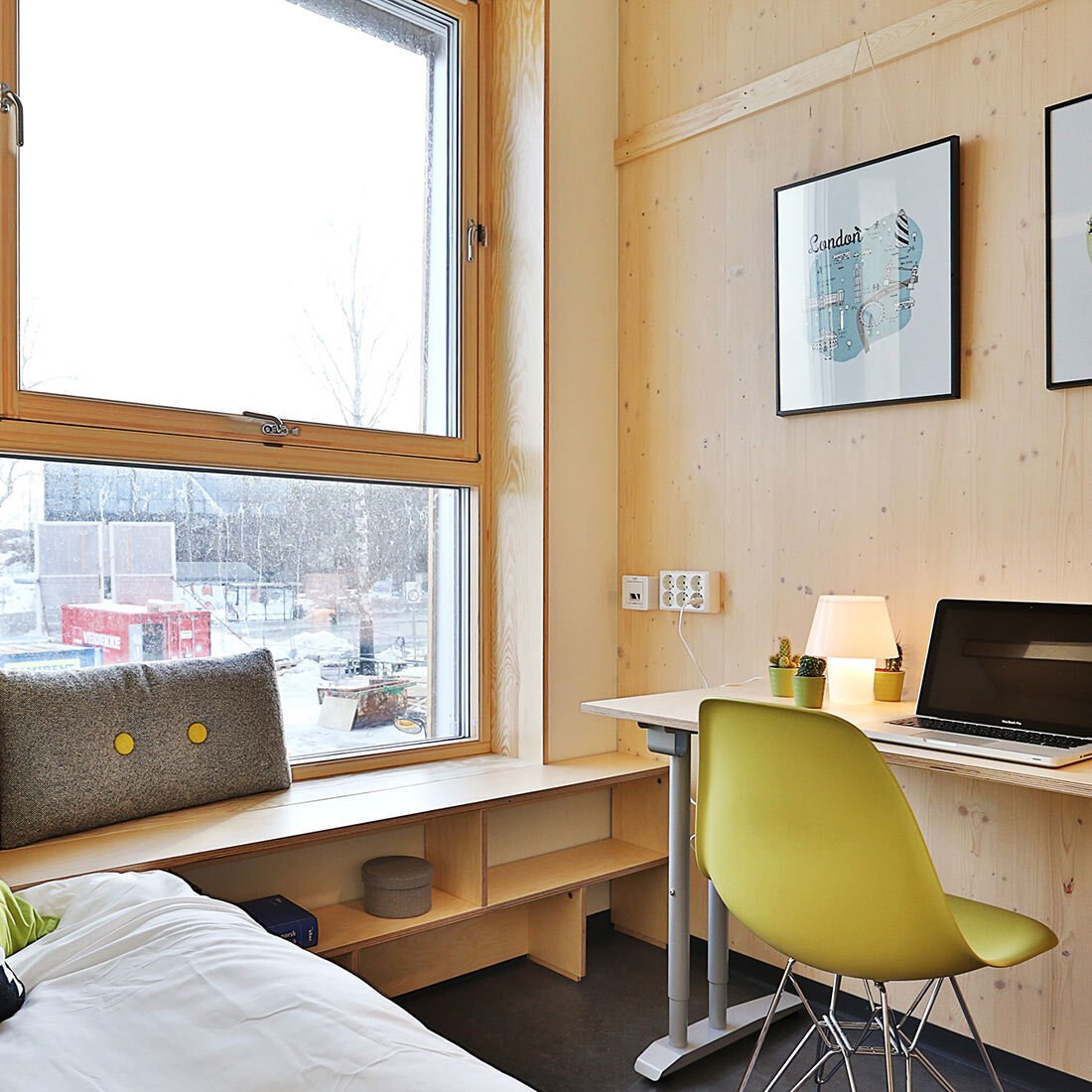 Interiør fra en hybel. Vi ser en seng, integrert hylle under et stort vindu, og en skrivepult med en gul stol inntil.