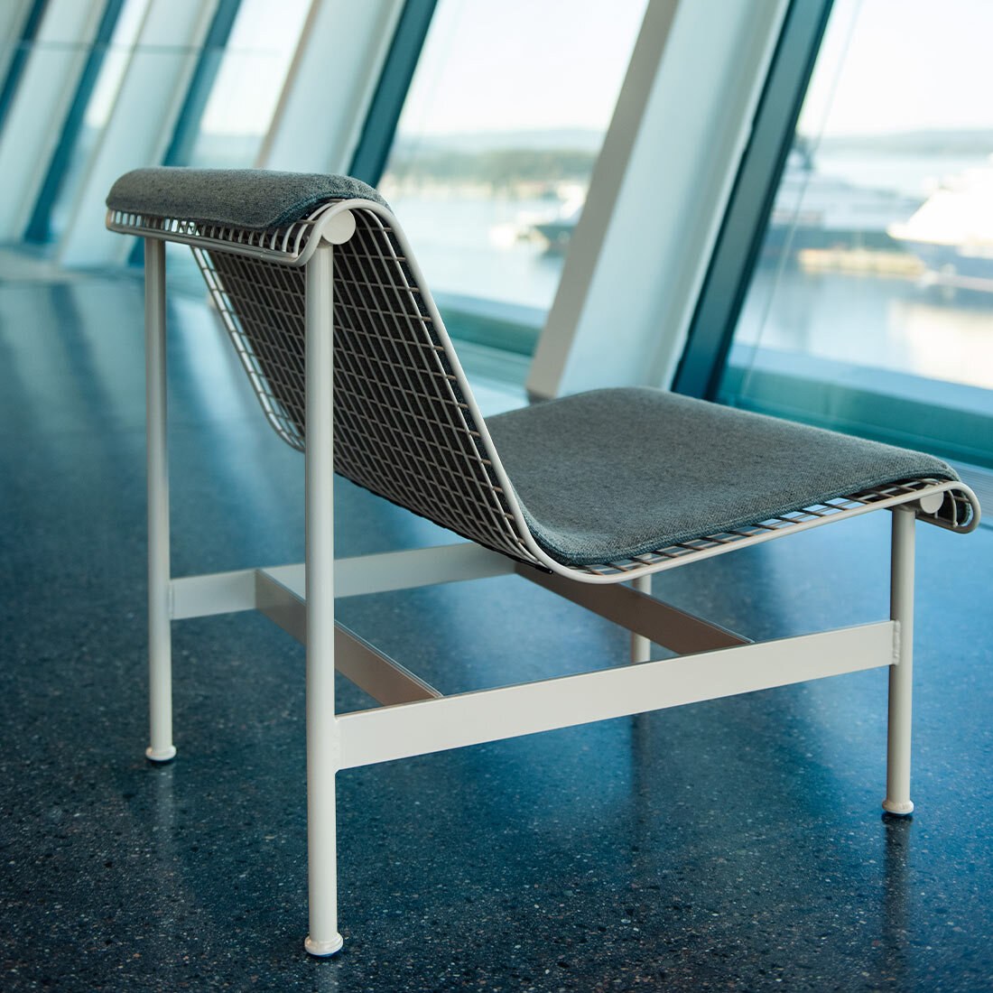 Et nærbilde av lounge-stolen. Den hat en kremfarget metallramme med nettinggitter som holder stolputen. Rammen har rette kanter, mens selve stolplaten har en avrundet form.