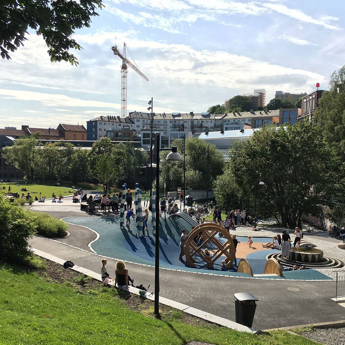 En park og et lekeområde ses fra en bakketopp. Voksne og barn sitter, står og leker. I randsonen av parken er grønne trær, og deretter tett bebyggelse.