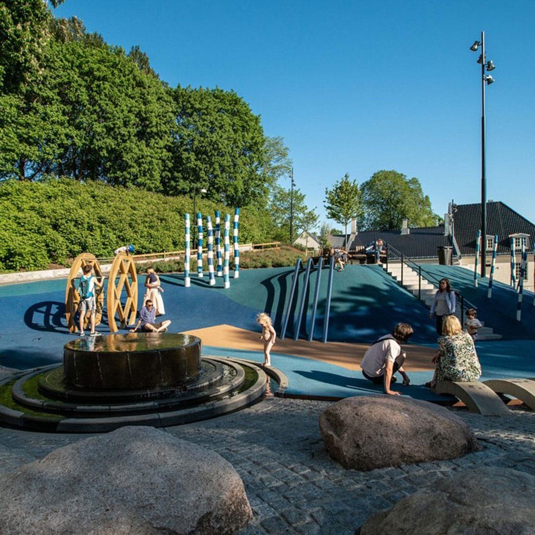 Barn leker på en lekeplass delvis dekket av blå gummi, delvis av brostein. Bakken følger terrenget, og lekeapparatene er utypiske: en fontene og vannkanal, et gult hjul, vinglete hvite og blå pinner som står opp av bakken, steiner og fire mørke blå rør som fører fra en liten bakketopp og ned.