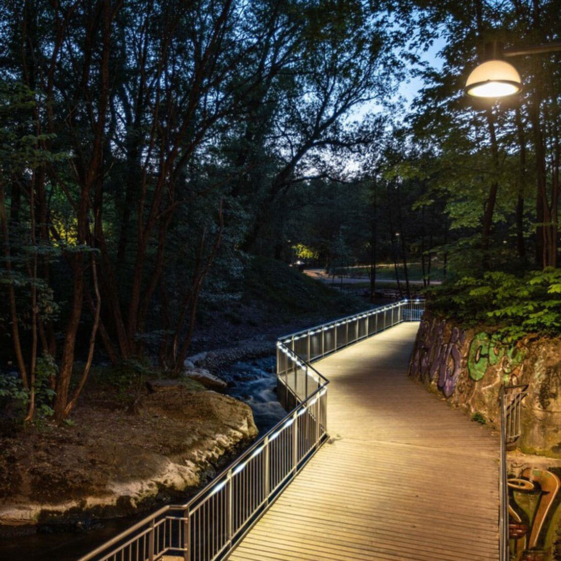 En gangbro følger en skrent langs en elv. Den en er opplyst av en gatelykt. Langs elvebredden er store trær.