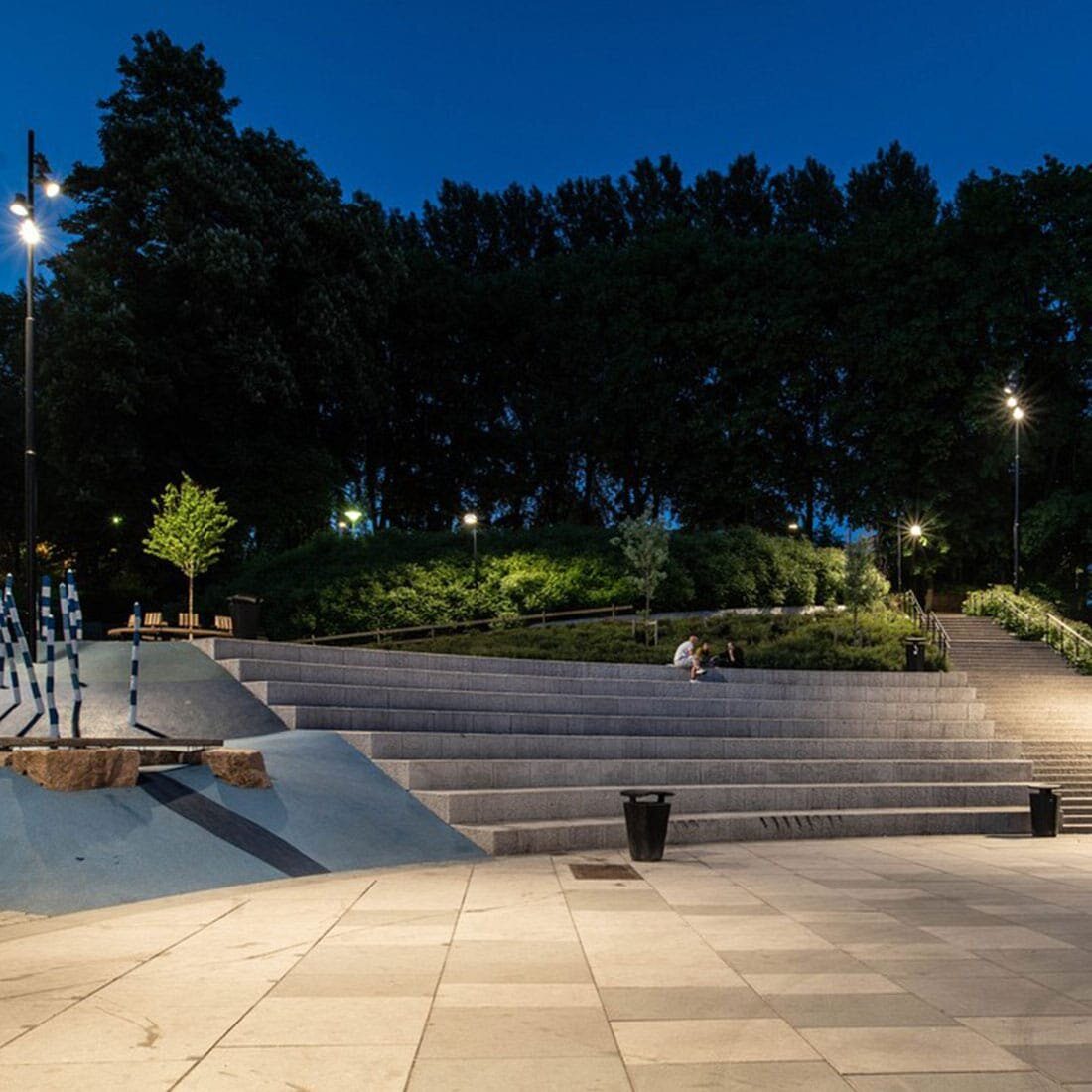 Vi ser opp på et sitteamfi i stein fra bakkeplan. Det er mørkt, gatelyktene er tent, og på toppen av amfiet sitter en liten gruppe mennesker. På bakketoppen bak amfiet ses buskbeplantning, og høye trær.