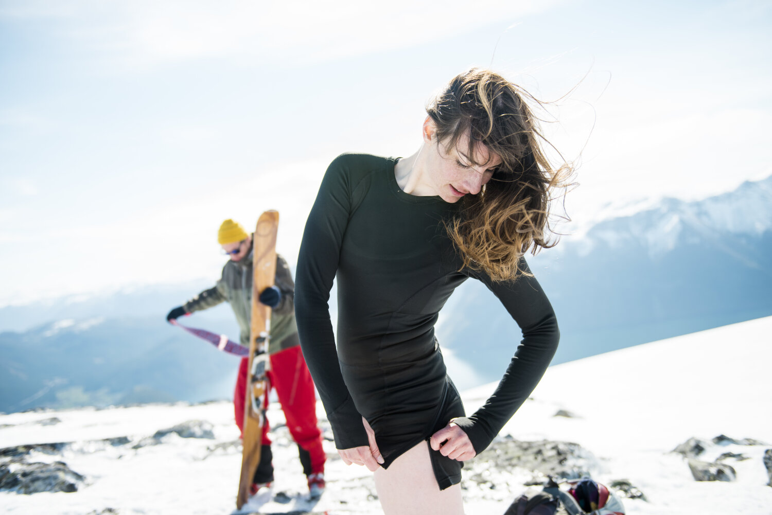 En kvinne på et snødekt fjell justerer en glidelås på en sort langermet body. Bak henne står en mann og tar en fell av en alpintski.