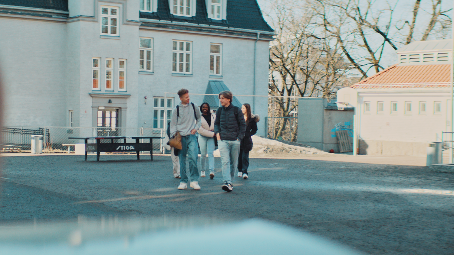 Fire ungdommer går på en asfaltert plass med skolebygninger i bakgrunnen.