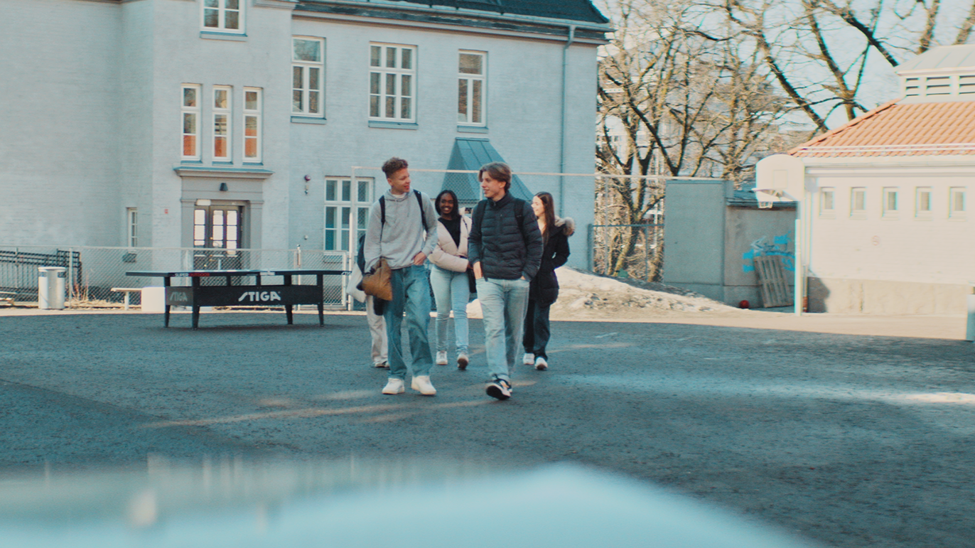 Fire ungdommer går sammen over en skolegård med skolen og et bordtennisbord i bakgrunnen.