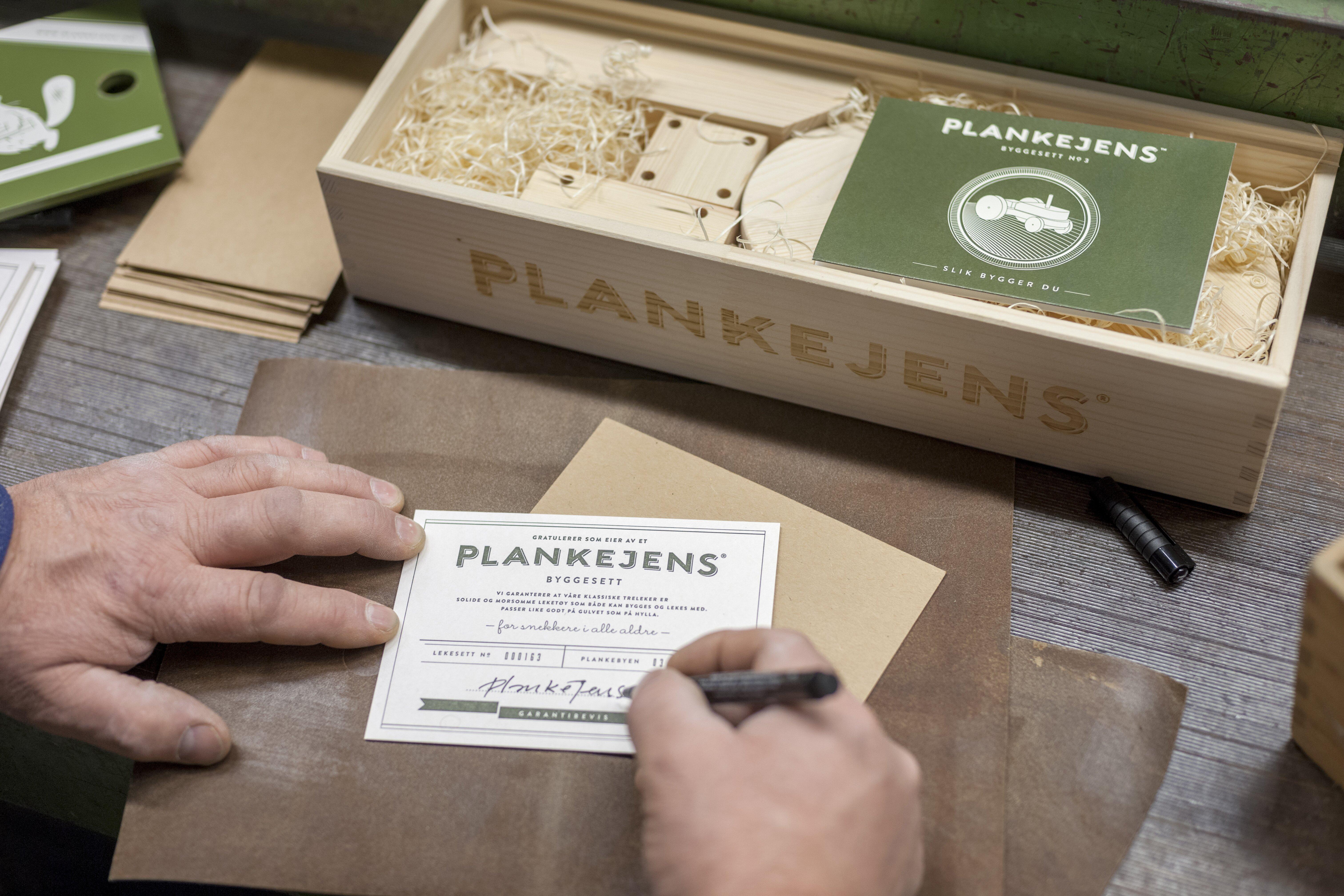 Fotografi av hender som signerer en etikett som tilfører et byggesett. Bak i bildet ser vi en trekasse med treklosser og trespon – et byggesett.