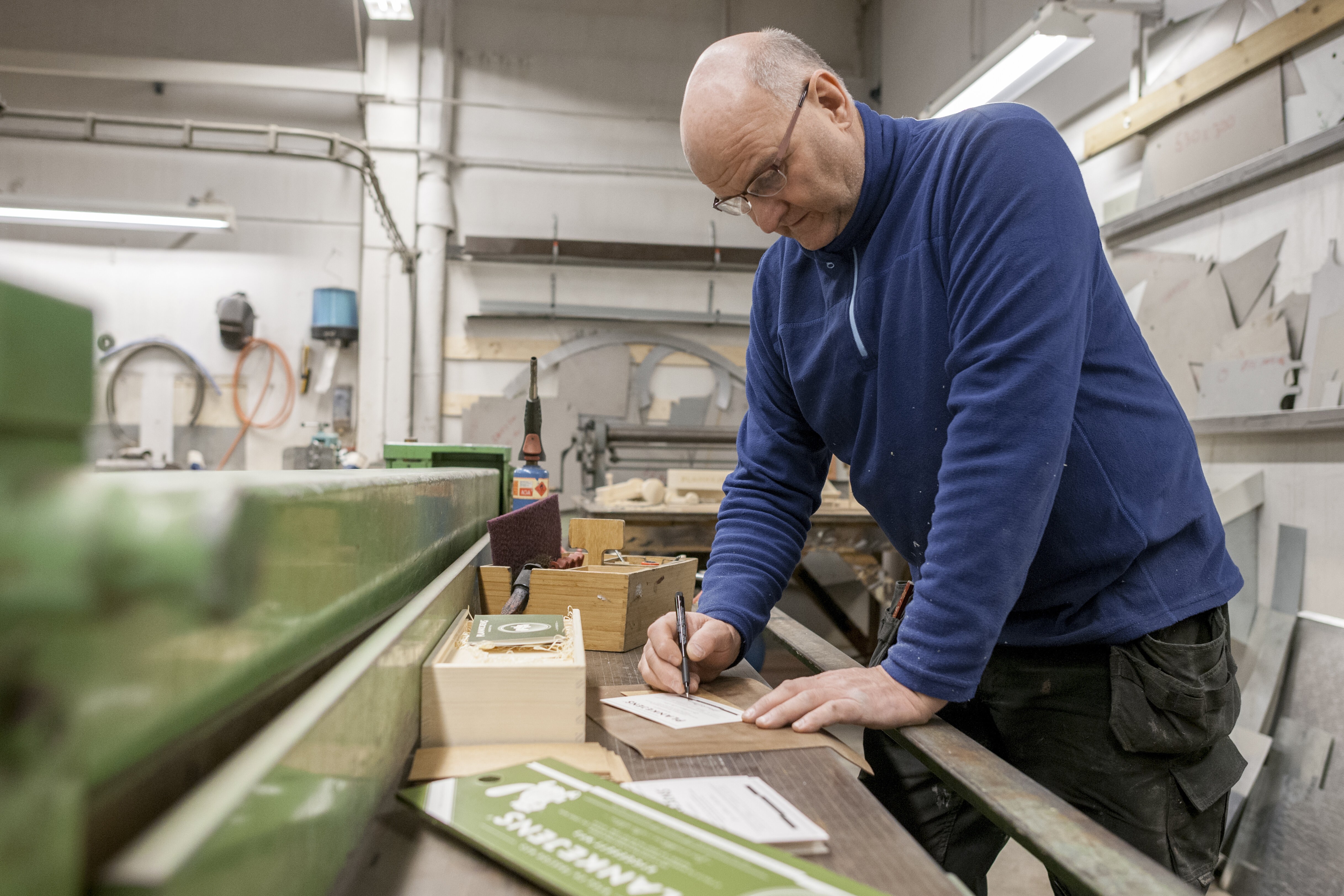 En mann står bøyd over en arbeidsbenk i et verksted. Han signerer en etikett til et byggesett.