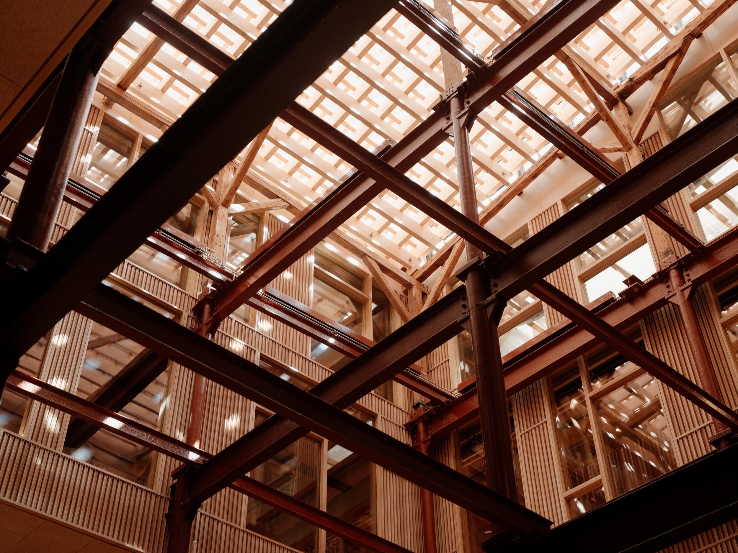 Interiør med synlige bærekonstruksjoner i tre og stål, store vindusflater og et lyst glasstak som slipper inn dagslys, sett nedenfra.