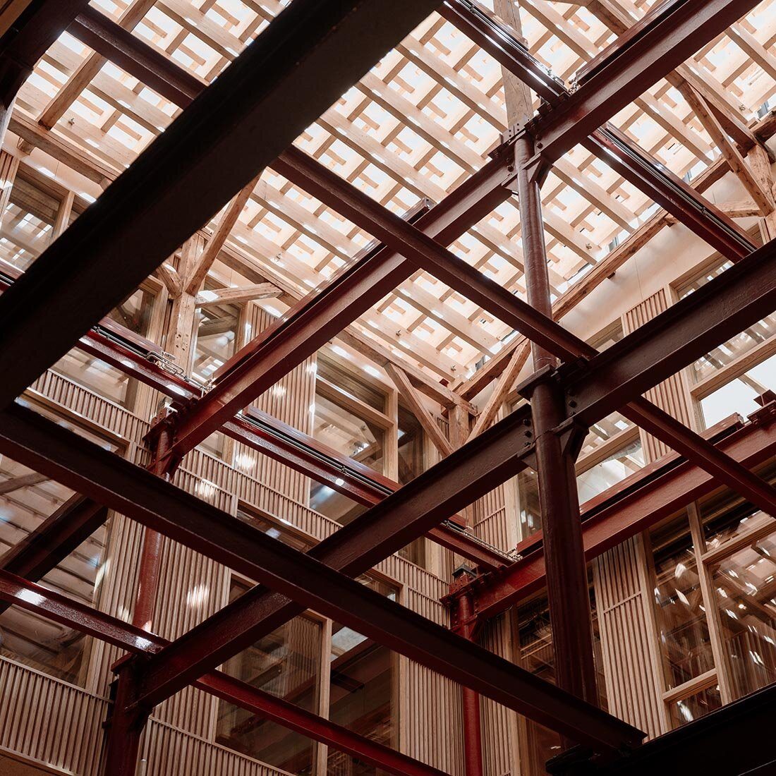 Interiør med synlige bærekonstruksjoner i tre og stål, store vindusflater og et lyst glasstak som slipper inn dagslys, sett nedenfra.