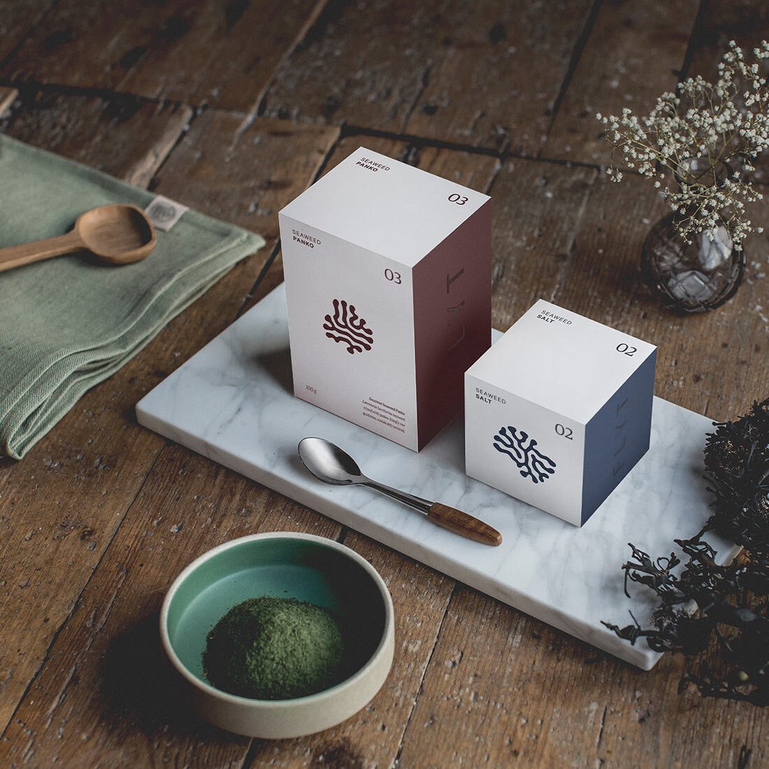 Bildet er et stilleben med to pappkartonger i ulike størrelser, emballasje for flyt, plassert på et marmorbrett på et rustikt trebord. Emballasjen har påtrykket hvert sitt merke, ulike varianter av et organisk, blæreaktig avtrykk.