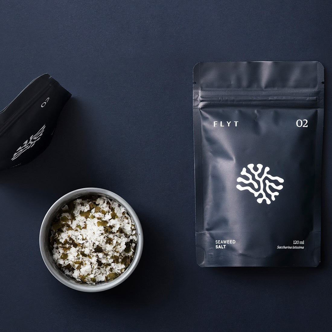 Bildet viser en plastemballasje for Flyt seaweed salt. Emballasjen er svart med hvitt trykk. Ved siden av emballasjen er en skål med grovt salt iblandet tangbiter. Bildet er tatt ovenfra.