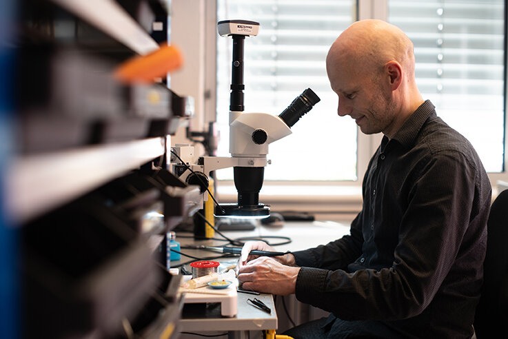 Mann sitter på kontor og ser i et mikroskop.