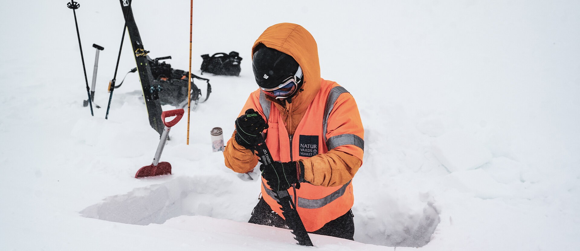 Bildet viser en person som står i dyp snø med en snøsag i hendene. i Bakgrunnen ski og staver.