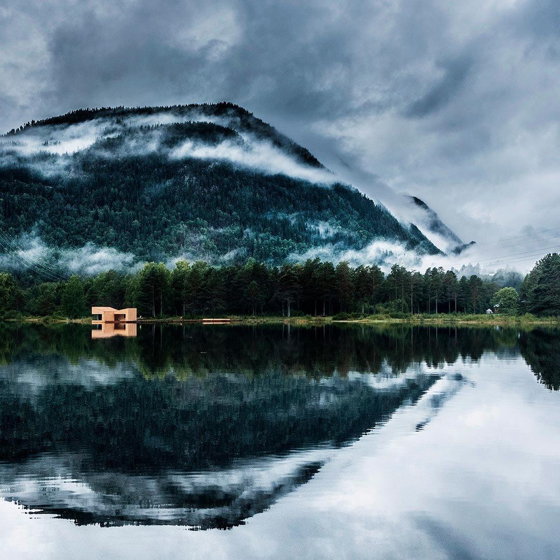 En badstu i lyst treverk er fotografert på avstand i et vakkert fjordlandskap, omkranset av skog og tåkedekte fjell. Fremst i bildet speiles landskapet i fjorden.