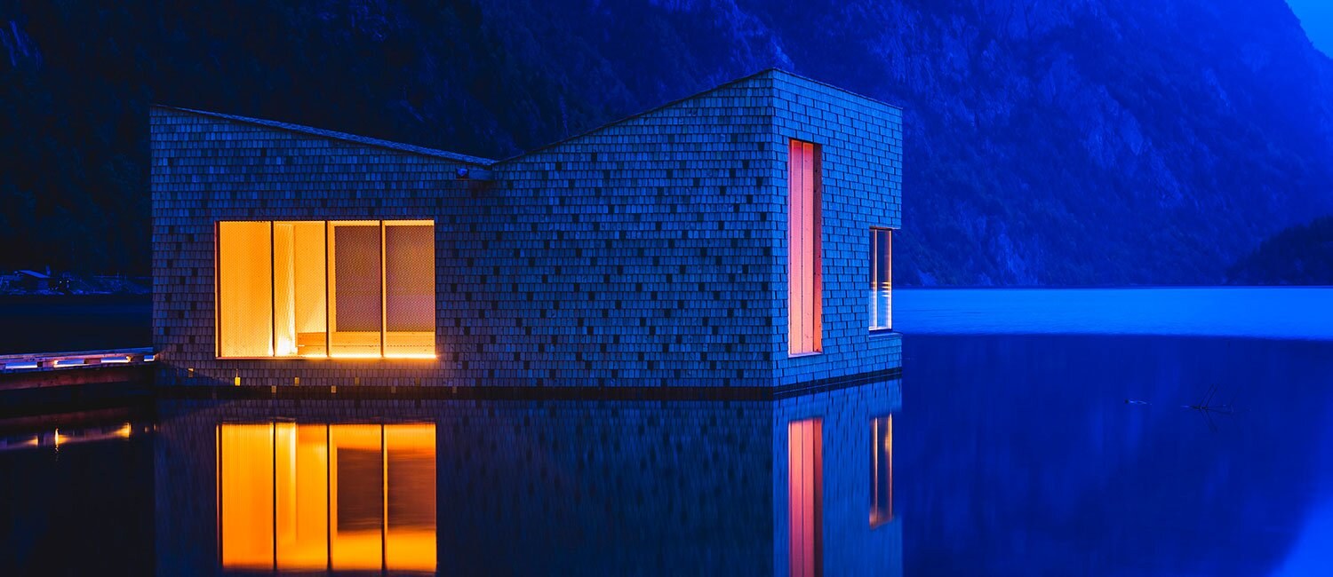 Bildet viser en badstu på en innsjø. Den er utformet i en karakteristisk form hvor taket vinkler oppover i hver ende. Omgivelsene bader i blått lys, mens innsiden er lyst opp i en varm gulfarge.