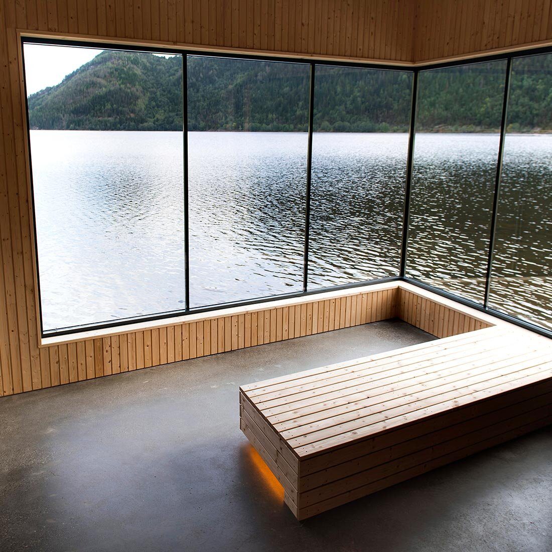 Interiørbilde fra badstuen. En integrert trekledt sittebenk er sentral i rommet, omgitt av vinduer fra gulv til tak med utsikt over fjord og skogdekte fjell.