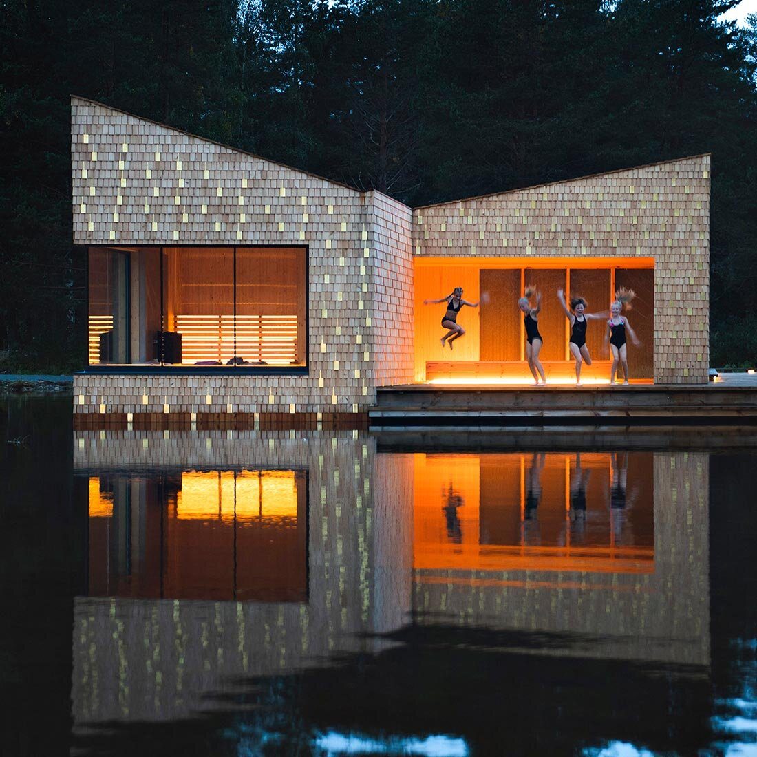 Et nærbilde av badstuen. Den er kledd i små treplater i ulike naturlige nyanser, og innimellom skinnende metallplater. Det er et stort vindu inn mot badstuen og et overbygg bak en badebrygge hvor fire unge jenter hopper ut i fjorden.
