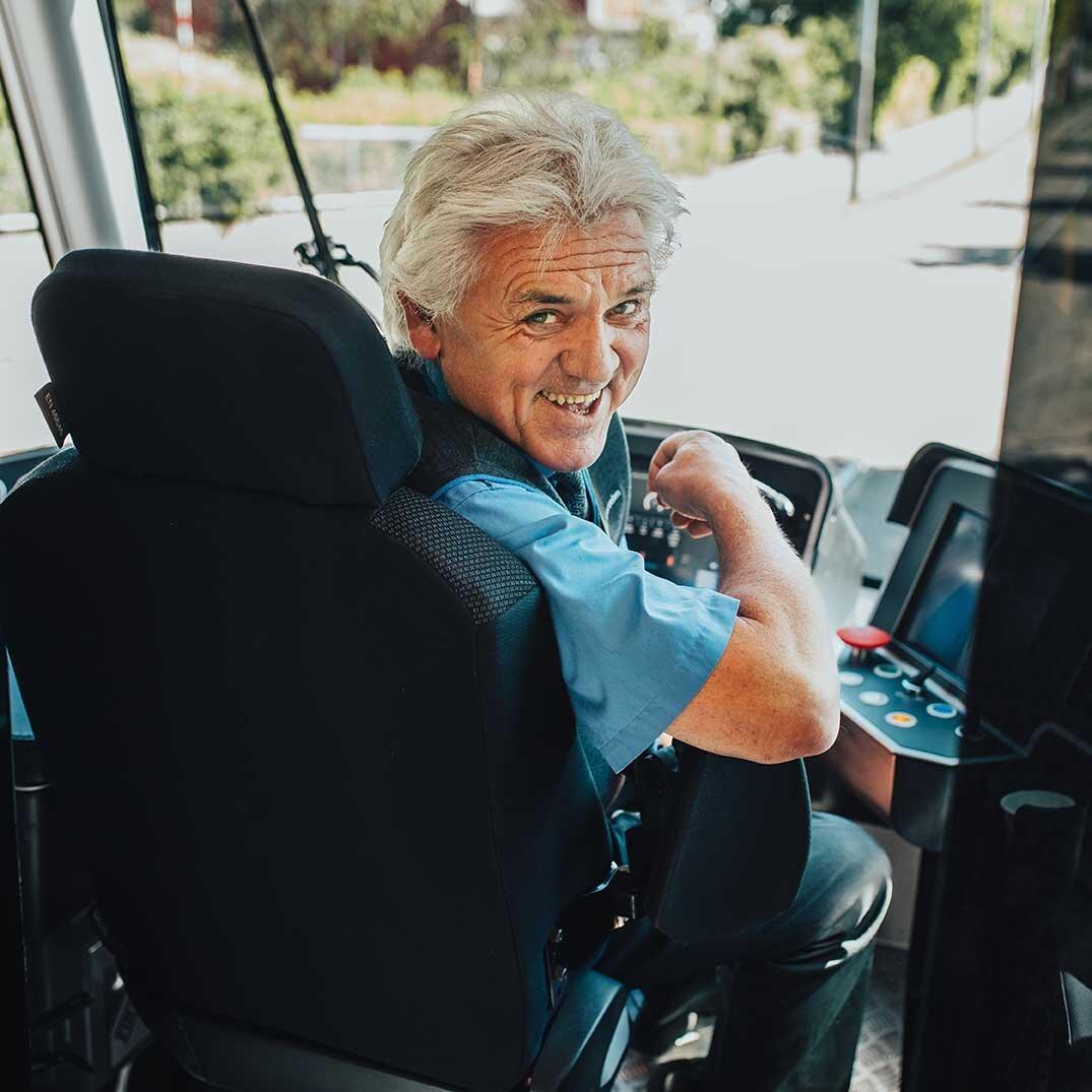 En trikkefører sittende i førersetet som snur seg bakover og smiler.