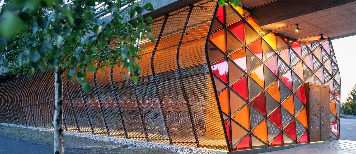 En konstruksjon under en bro. Konstruksjonen har et skjelett i metall. Kortsiden ha ren dobbel dør, og er dekt i glasstrekanter i rødt, oransje og gult. Langsiden er kledd i netting.