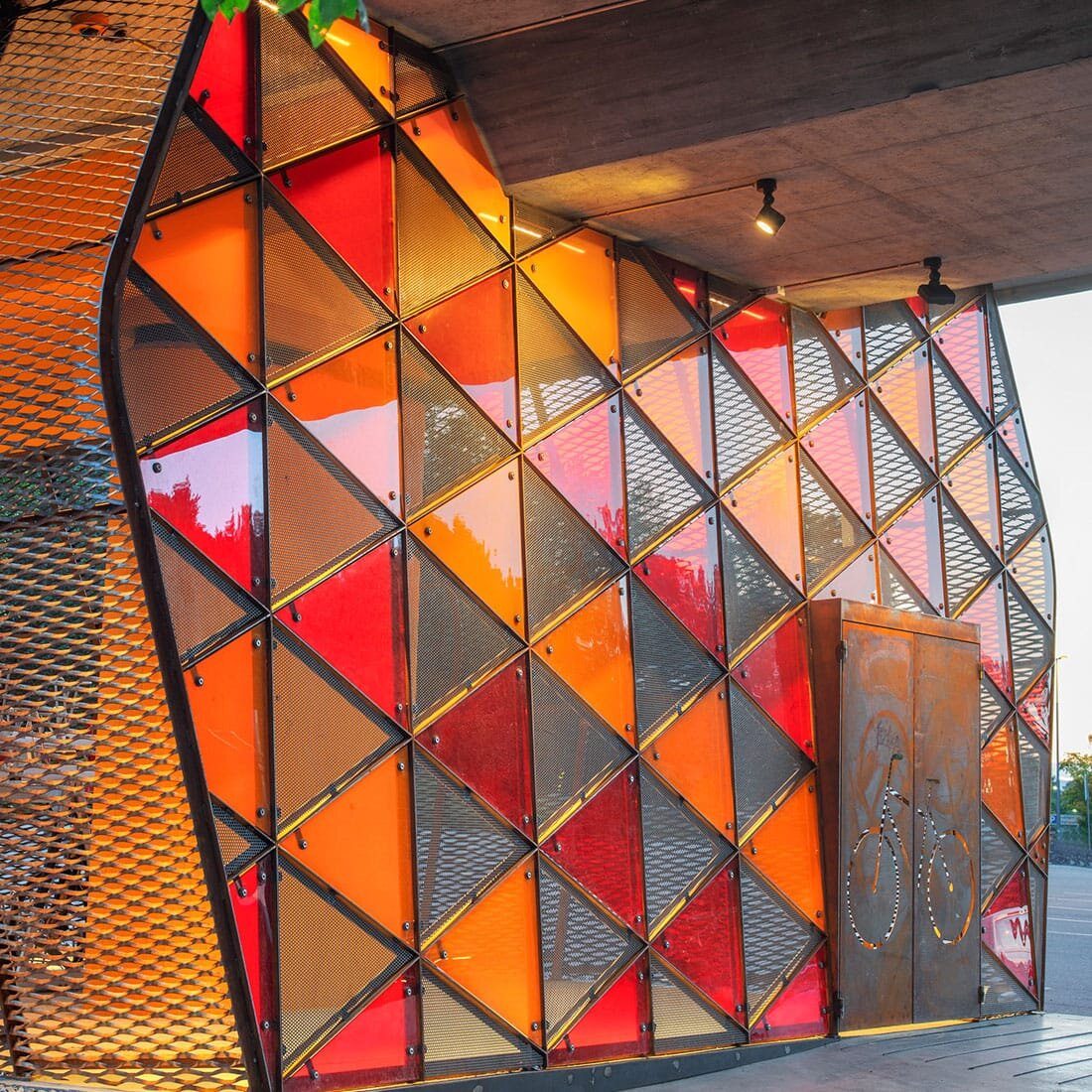 Et nærbilde av konstruksjonen som viser skinnende innfelte glasstrekanter i fasaden, og kontrast mot betong i brotaket og døren i grovt metall.