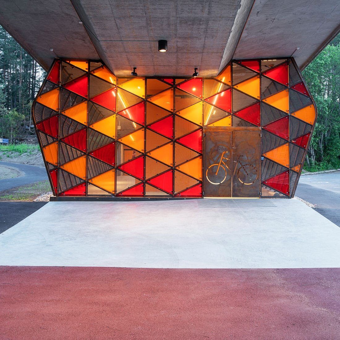 Vi ser kortsiden av en konstruksjon plassert under en bro. Den har en dobbel dør prydet av en innrisset sykkel. Fasaden er dekt i glasstrekanter i rødt, oransje og gult.