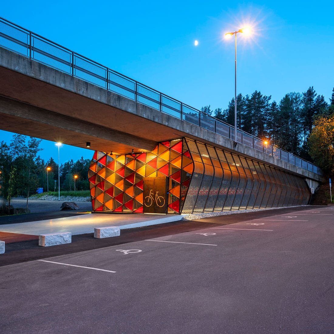 En konstruksjon under en bro. Konstruksjonen har et skjelett i metall. Kortsiden ha ren dobbel dør, og er dekt i glasstrekanter i rødt, oransje og gult. Langsiden er kledd i netting.