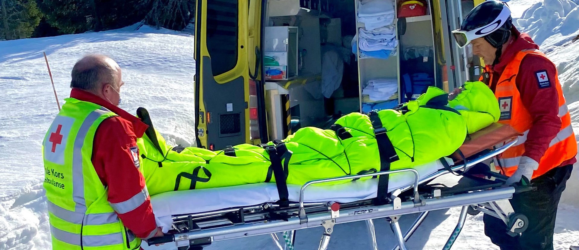 Bildet viser to redningsarbeidere som løfter en båre med en pasient inn i en ambulanse. Pasienten er pakket inn i en signalgul isolasjonspose.