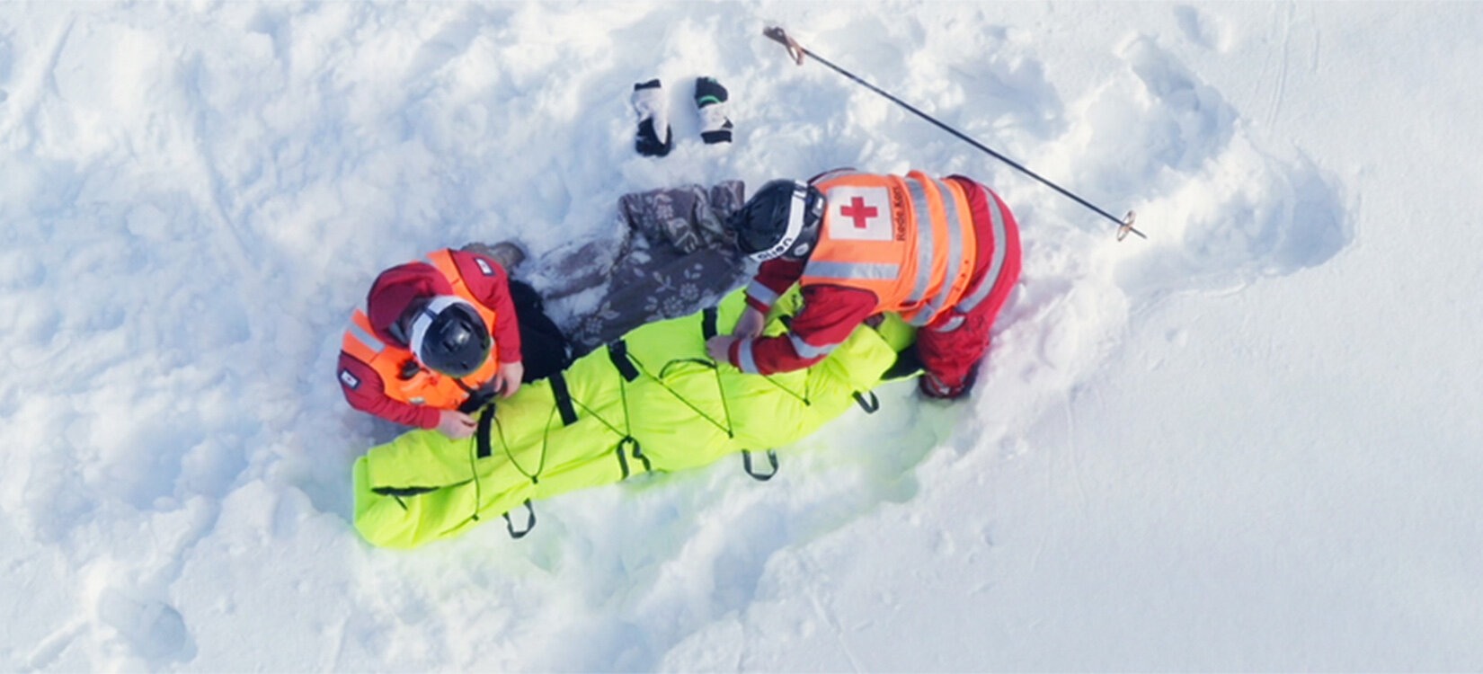 Foto av to redningsarbeidere i snøen som arbeide med å pakke en pasient inn i en isolasjonspose. Bildet er tatt i fugleperspektiv.