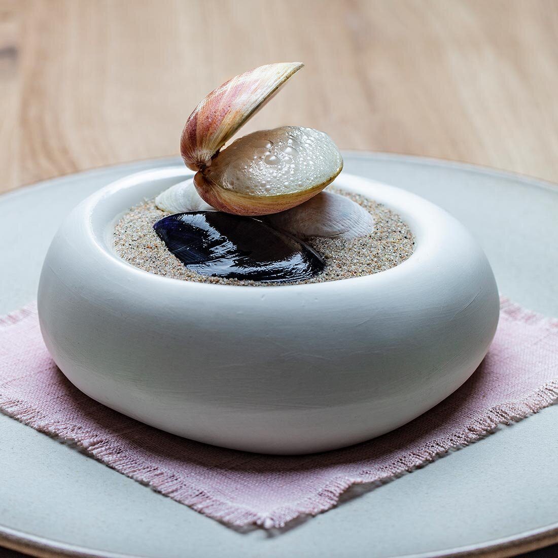Foto av en nøye presentert matrett dandert i en skål formet som en nedsunket bolle. Et skjell er åpent og fylt med skum, og plassert på toppen av tre skjell som ligger i sand.