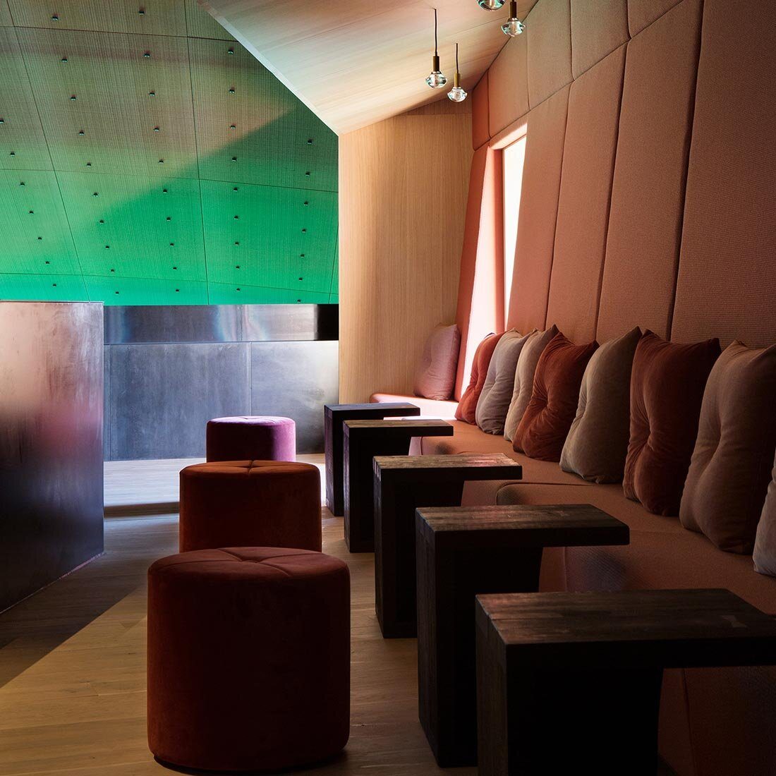 Interiørfoto av Under. En sofabenk med puter i nyanser av burgunder, rosa og grå. Småbord og høye sittepuffer. En vegg er polstret, taket er i eikefiner, og en annen vegg ser ut til å være kledd i metallplater.