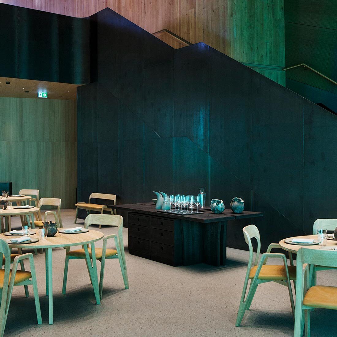 Interiørfoto av spisesal. En svartkledd trapp leder ned fra etasjen over og dannet bakgrunnen i bildet. Flere bordsettinger av tremøbler på varmt grått gulv.