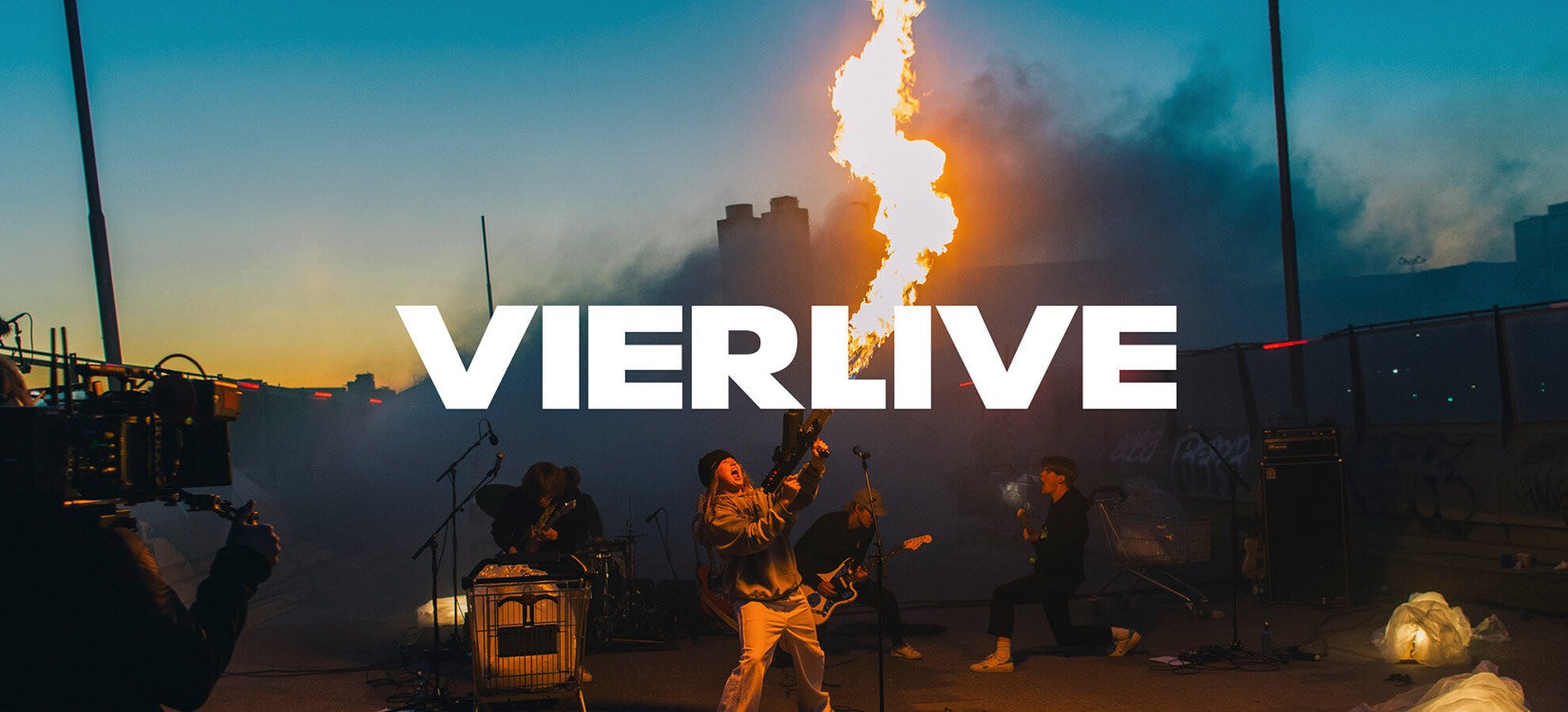 Et utendørs sceneshow med en person som holder en flammekaster. Over bildet står: "VIERLIVE"