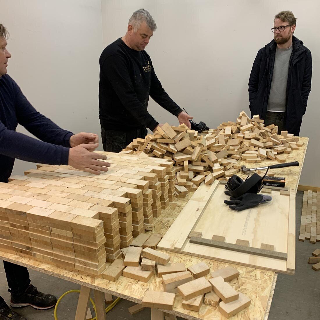 Tre menn står rundt et bord fylt av trekubber. Noen ligger løst i en haug, mens andre er satt sammen til større plater av kubber.
