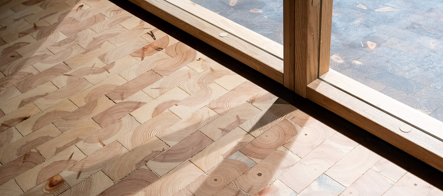 Utsnitt av kubbegulv hvor sollyset treffer fra vindu som går helt ned til gulvet. Det er tydelig spill i treverket, med årringer og fargevariasjoner.