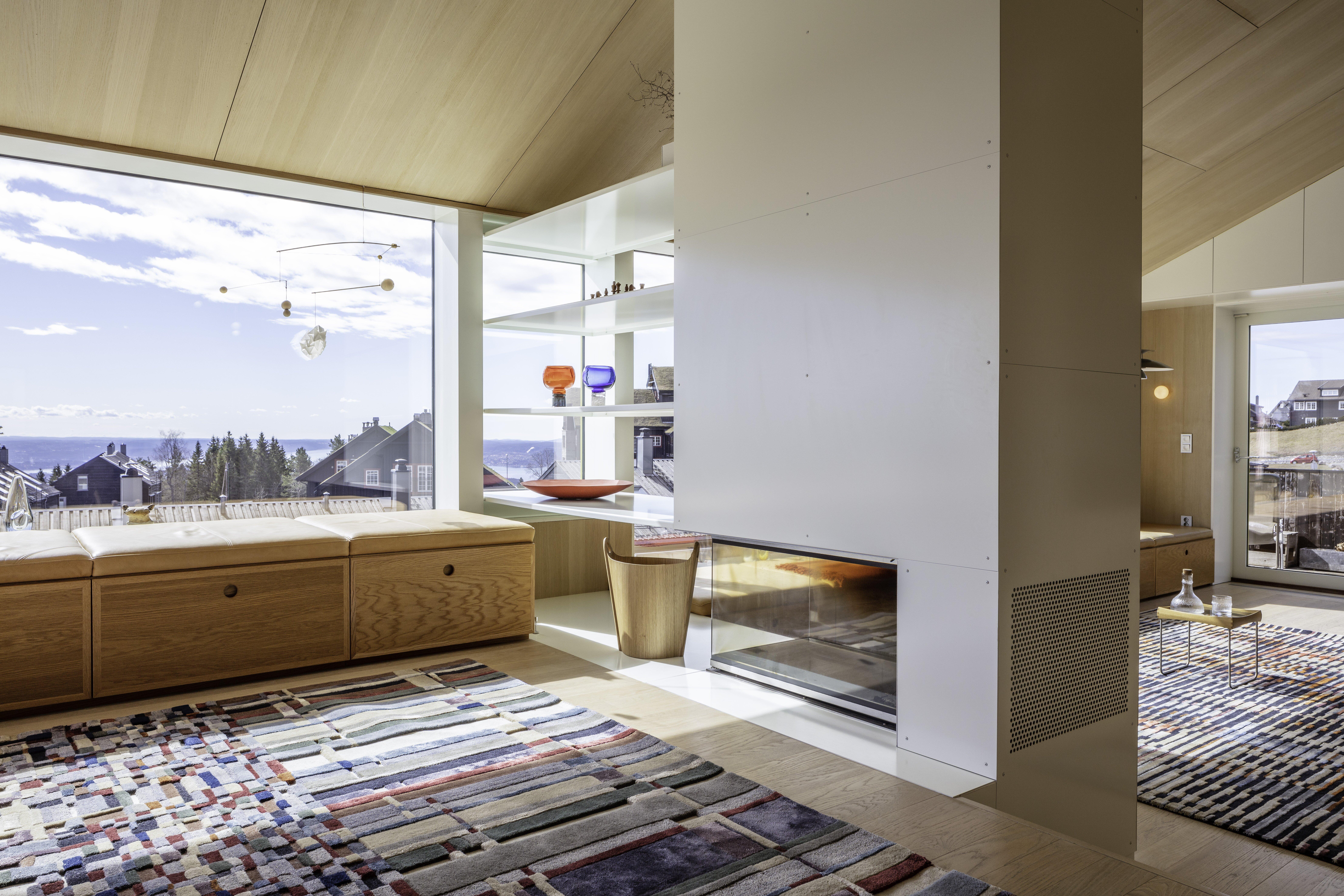 Et interiørfoto av en moderne innredet stue, men naturlige treoverflater og innfelt peis. På gulvet ligger et vevet teppe i stripete og rutete mønster i dempede nyanser av grønt, blått og rødt.