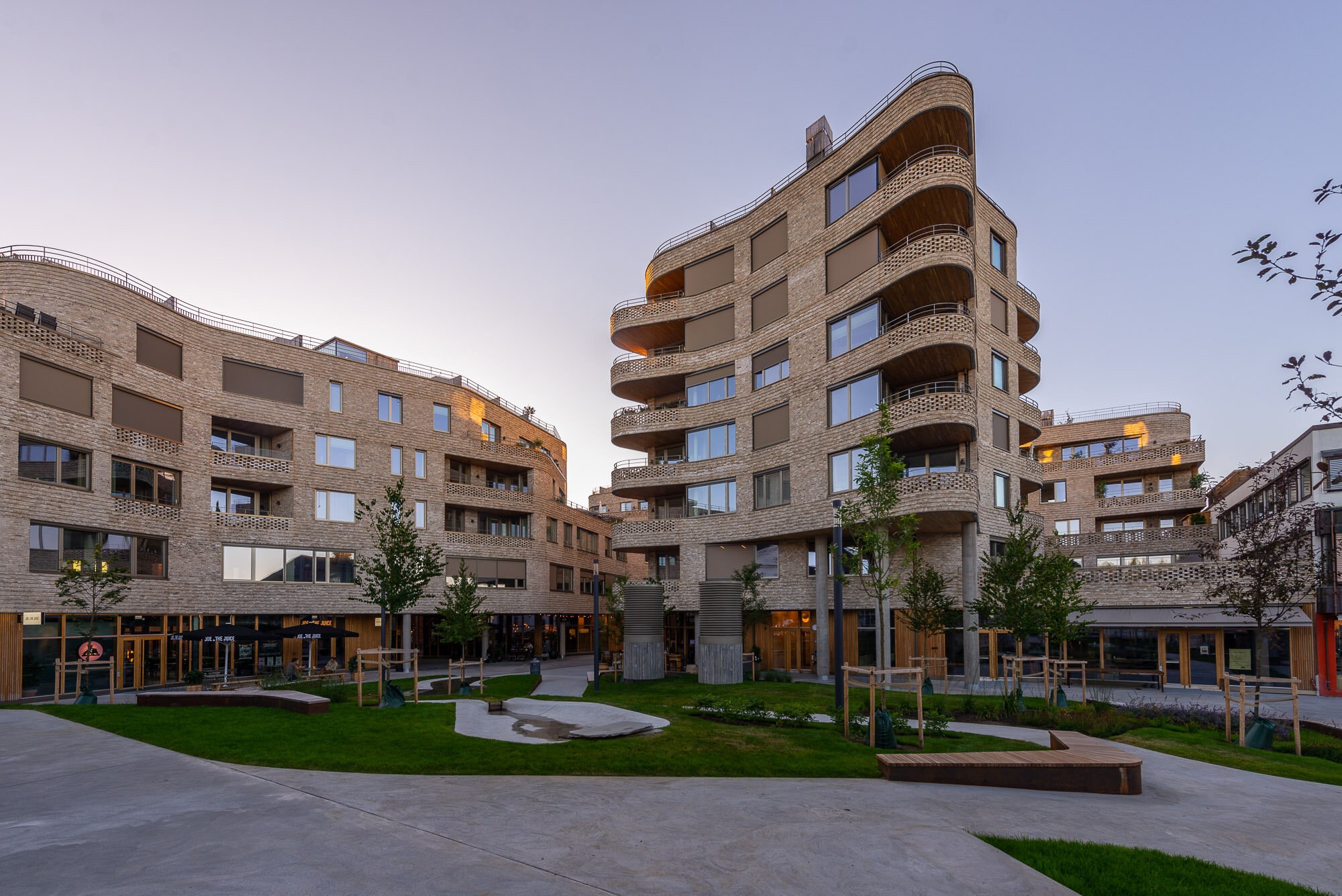 Foto av et urbant byrom innrammet av buende blokkbebyggelse over 5–7 etasjer med teglsteinsfasader. Utadrettede førsteetasjer åpner opp mot et byrom med beplantning, benker og gangveier.
