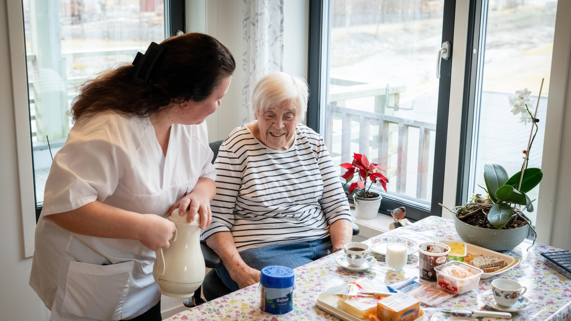 En hjelpepleier serverer kaffe til en eldre kvinne.