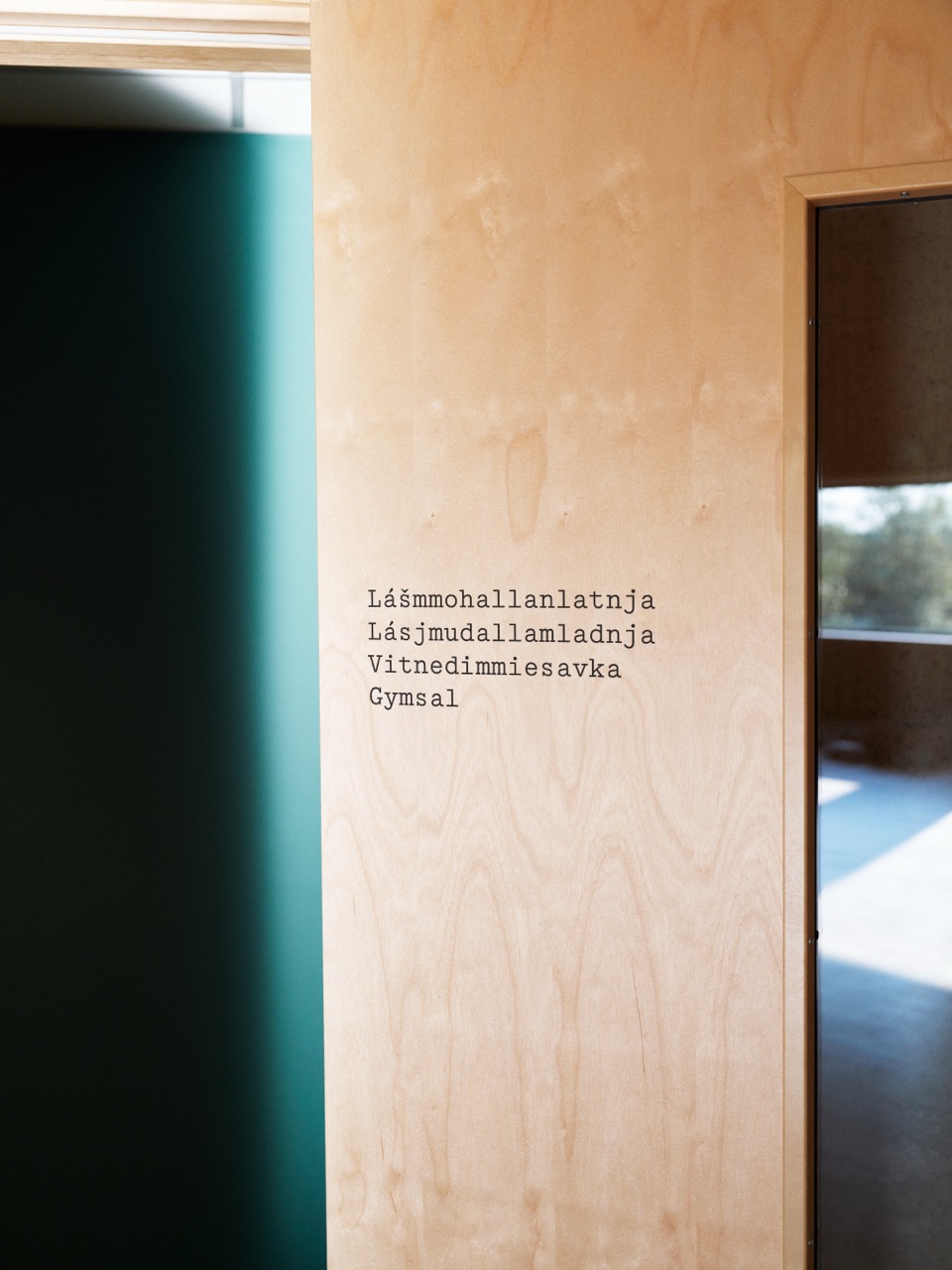 Nærbilde av skilt i trefinér med ordet gymsal i sorte bokstaver på fire språk.