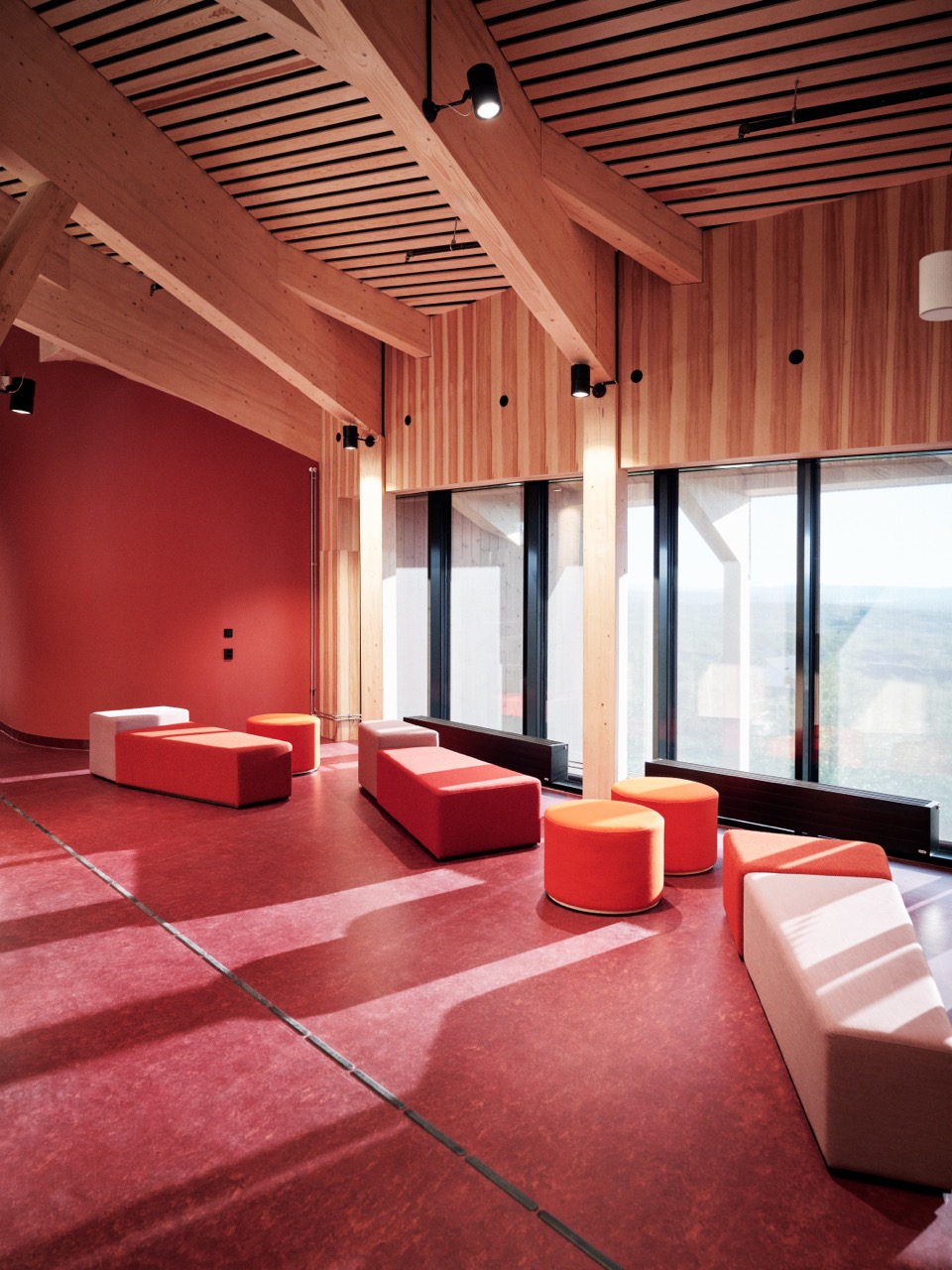 Interiør i et moderne rom med store, synlige trebjelker og panelte vegger. Rommet har rødt gulv, lave sittemøbler i røde og lyse farger og store vinduer som slipper inn mye dagslys.