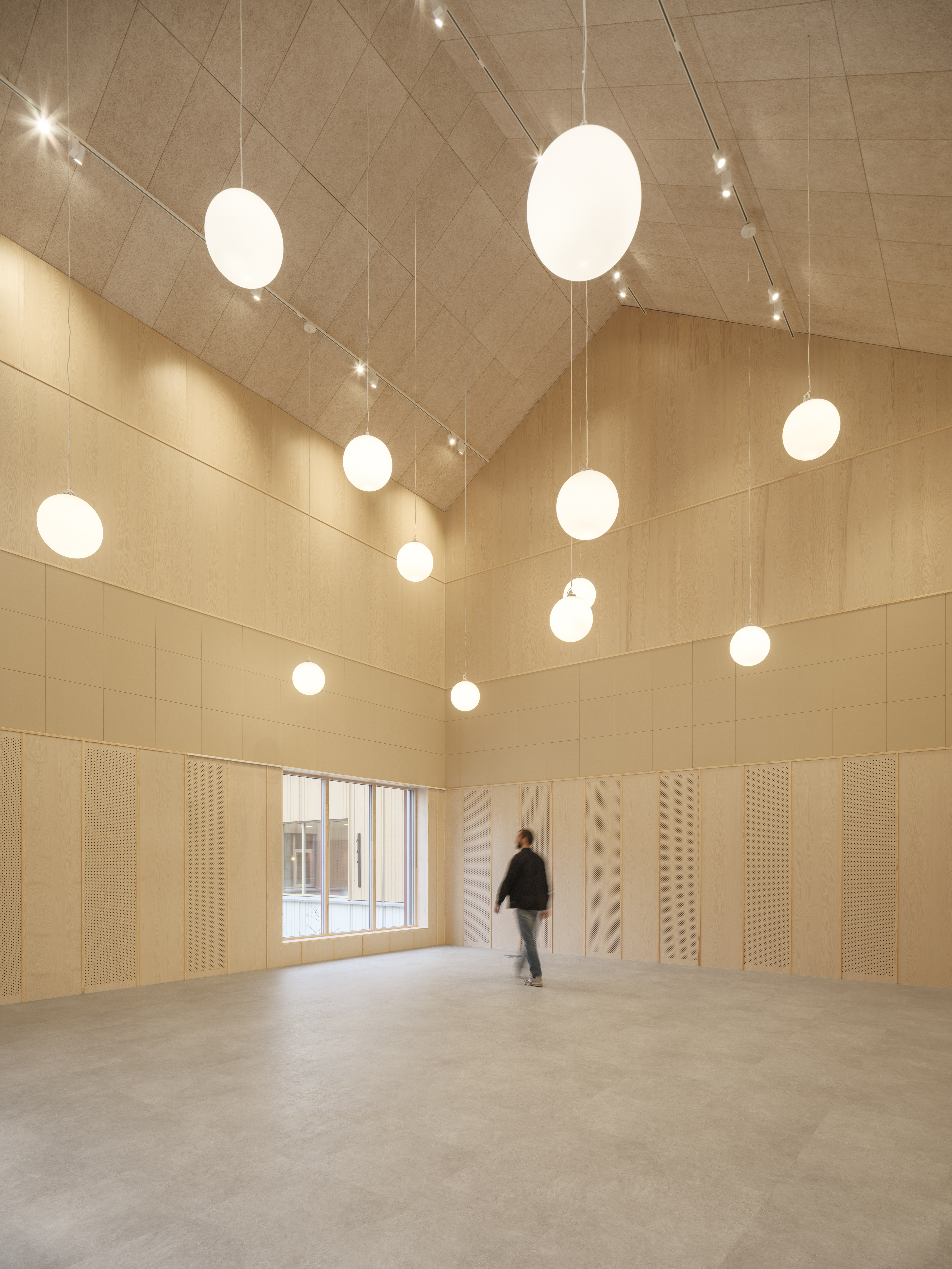 Et stort lyst rom uten møbler men med runde lamper hengende ned fra taket. En mann går mot døren.