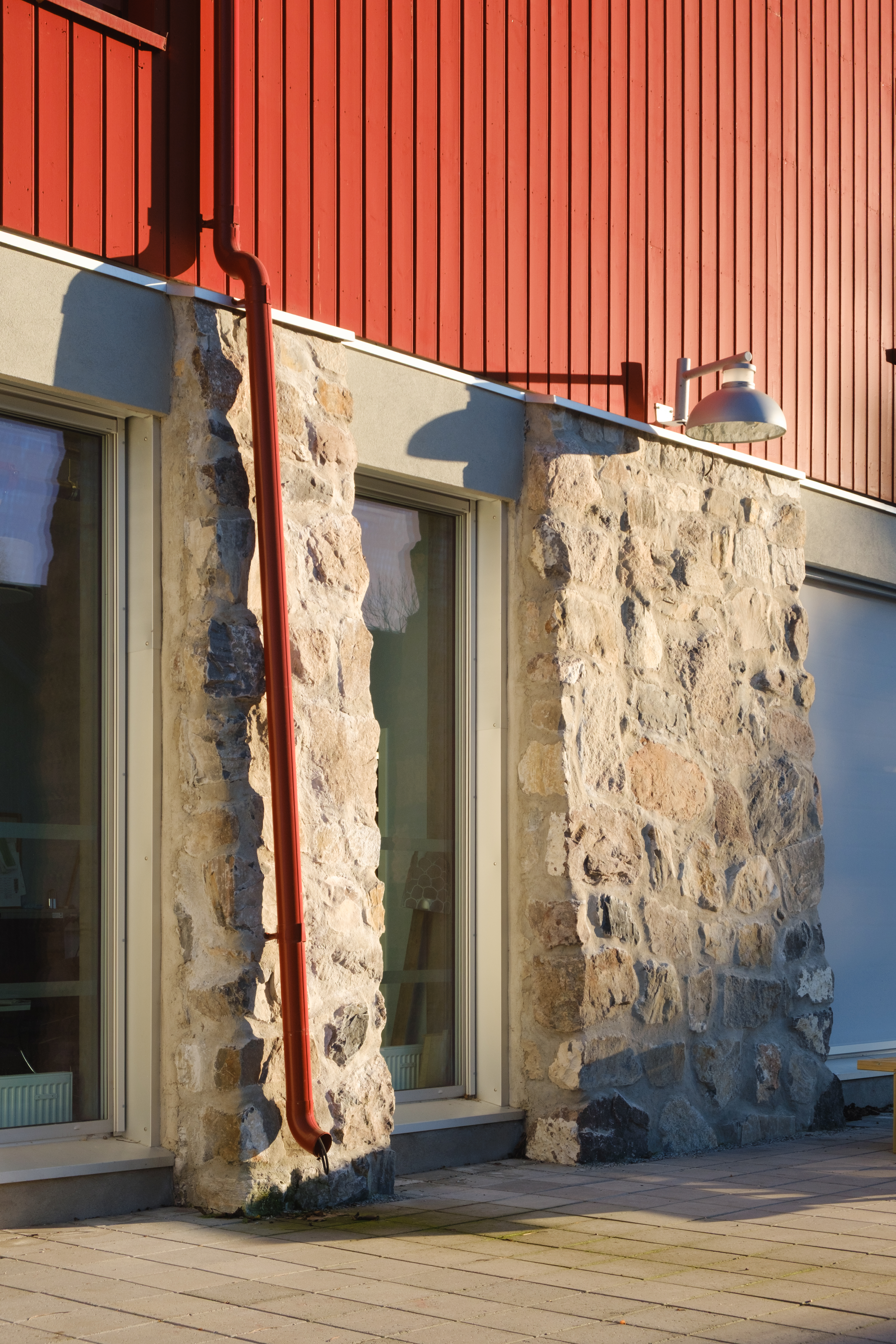 En grunnmur av naturstein med en rød panelt vegg over. Et rødt taknedløp går ned langs veggen og muren mot bakken.