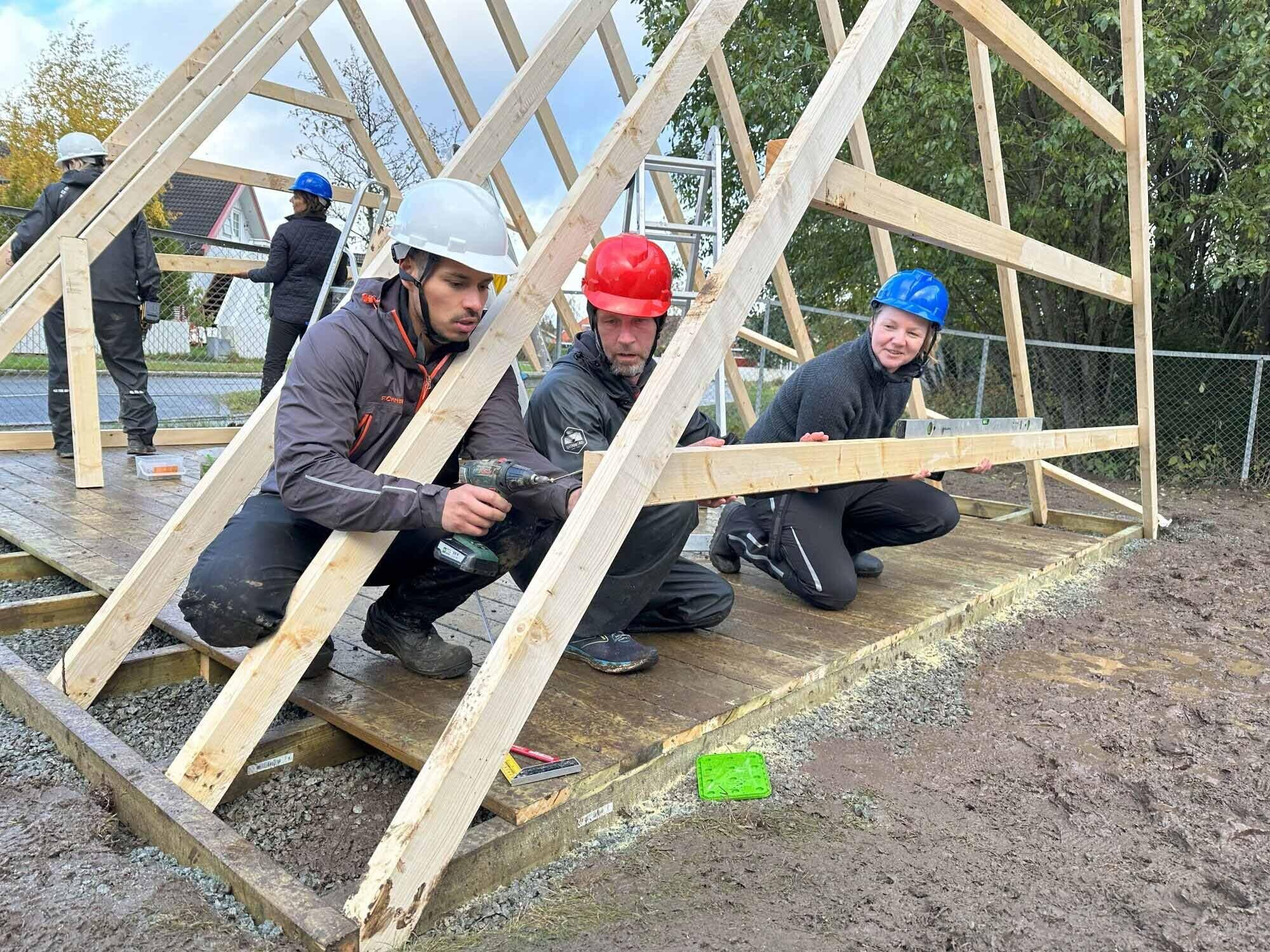 Flere folk med hjelm som setter opp et reisverk til et lite bygg sammen på dugnad.
